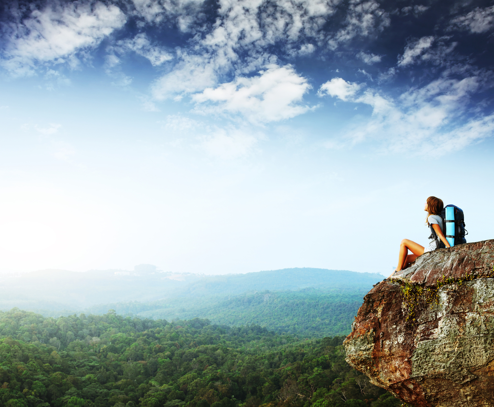 Young woman with backpack sitting on cliff and looking to a sky.