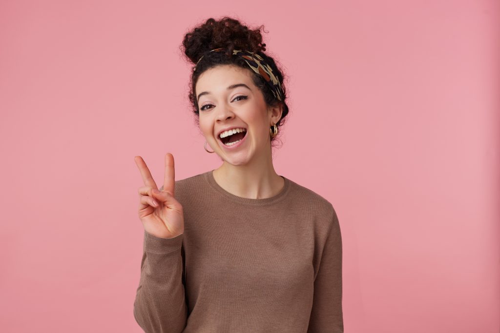 Cheerful woman, beautiful girl with dark curly hair bun. Wearing headband, earrings and brown sweater. Has make up. Showing peace sign. Watching at the camera isolated over pastel pink background