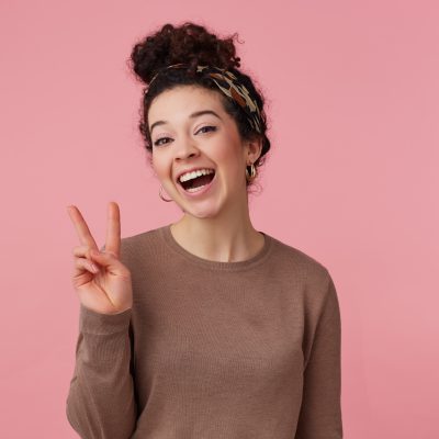Cheerful woman, beautiful girl with dark curly hair bun. Wearing headband, earrings and brown sweater. Has make up. Showing peace sign. Watching at the camera isolated over pastel pink background