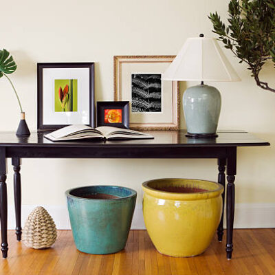 Home interior living room decorating arrangement featuring a classic, traditional wooden side table with arrangement of a celadon ceramic lamp, flower and plant photographs, open book, potted basil tree in large pot, and other color coordinated pottery on the hardwood floor. An urban scene showcase for style and design.