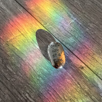 A small stone engraved with the words “one day at a time,” resting on wood as rainbow light falls across it.