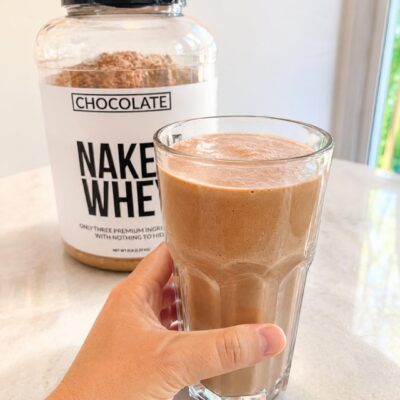 Creamy chocolate protein shake made with Naked Whey chocolate protein powder in a clear glass on a bright kitchen countertop.