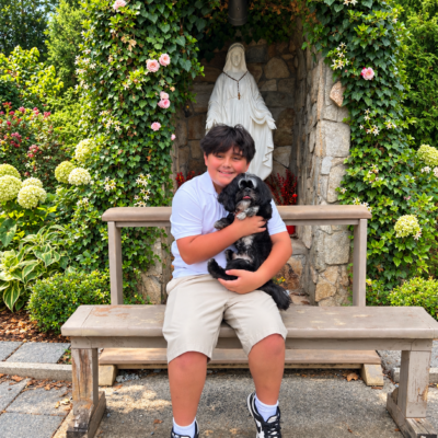 Joaquín sits on a wooden bench holding his black and white Shih Tzu, Panda, in front of a Virgin Mary statue surrounded by flowering vines and greenery.